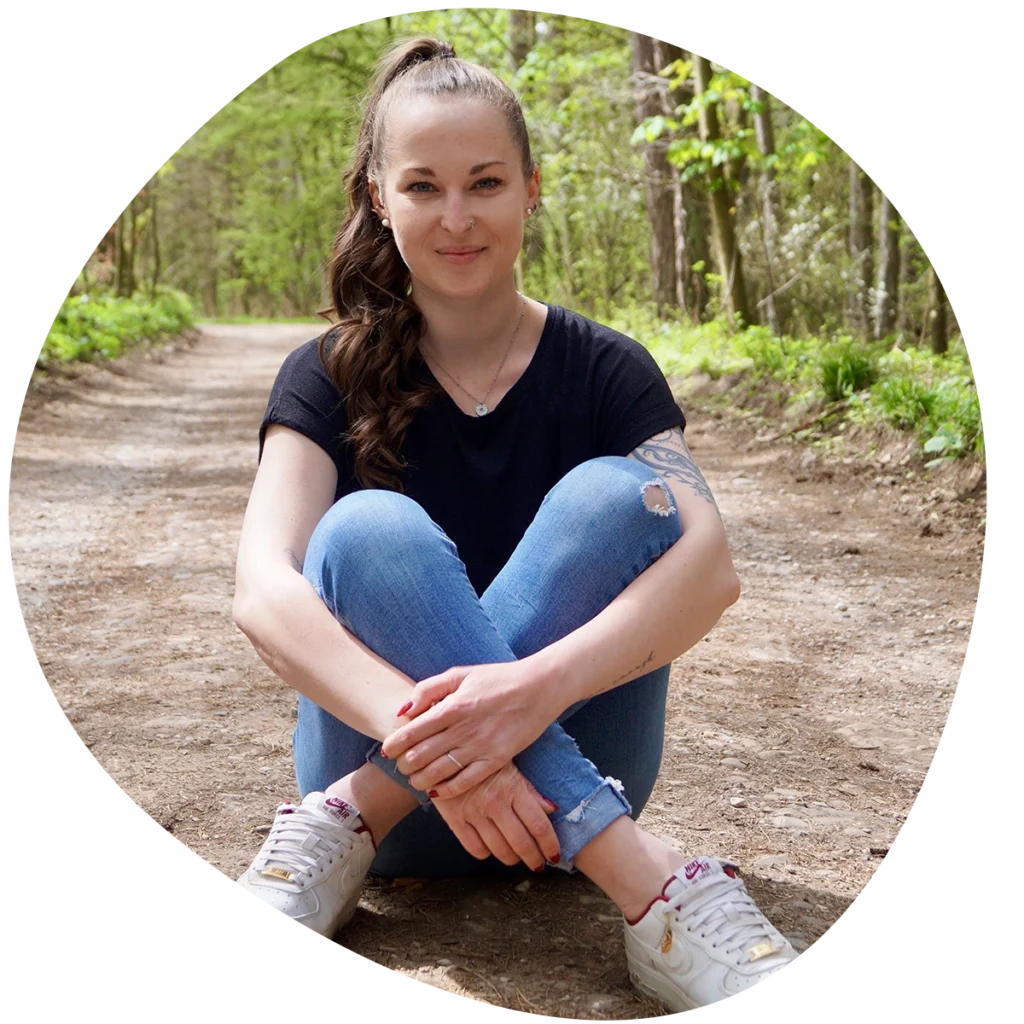 Sandra Teuchert sitzt auf dem Waldboden mit verschränkten Armen und Beinen, trägt blaue Jeans und weiße Sneakers.